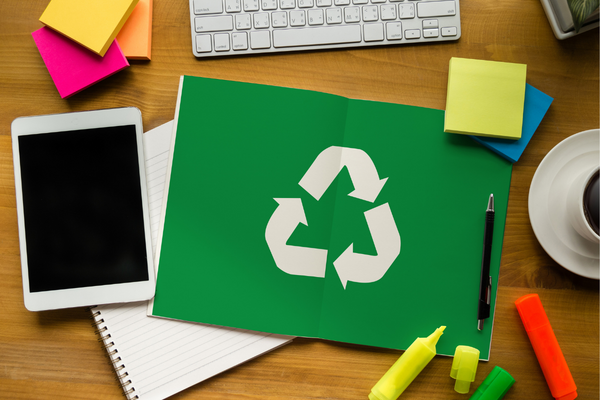A desk with post-it notes, an iPad, and a green open notebook with the recycling symbol printed across two pages
