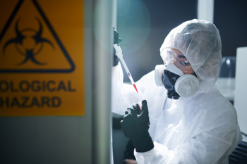 A woman in PPE holding a test tube and chemicals in a pipette in front of a biological hazard warning sign