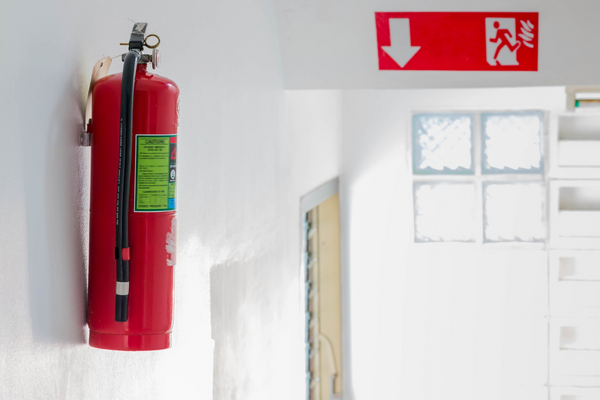 A white corridor with a bright red Fire Exit sign overhead and a red fire extinguisher attached to the wall on the left